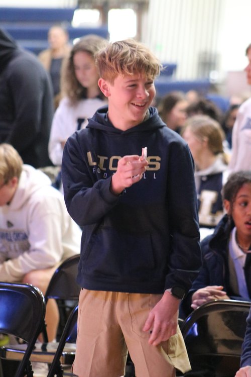 A young boy with curly hair, wearing a dark hoodie and khaki pants, smiles and gestures playfully while standing among a group of seated peers in a gymnasium.