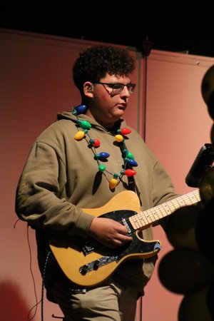 A young musician wearing a hoodie and glasses plays an electric guitar while adorned with a colorful string of lights around his neck, set against a softly lit background.