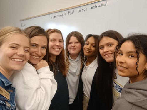 A group of seven young women smiles together in a classroom setting, with a whiteboard in the background that has text written on it.