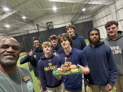 A group of seven young men and a coach pose together in an indoor sports facility, with one of the young men holding a tray of donuts and a cake with candles, celebrating a birthday.