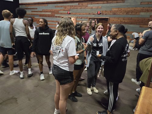 A group of young people, some in athletic wear, are gathered in a casual setting, engaging in conversation and socializing with smiles and laughter.