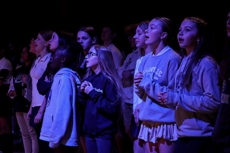 A group of young people stands together, singing with focused expressions, illuminated by soft purple lighting.