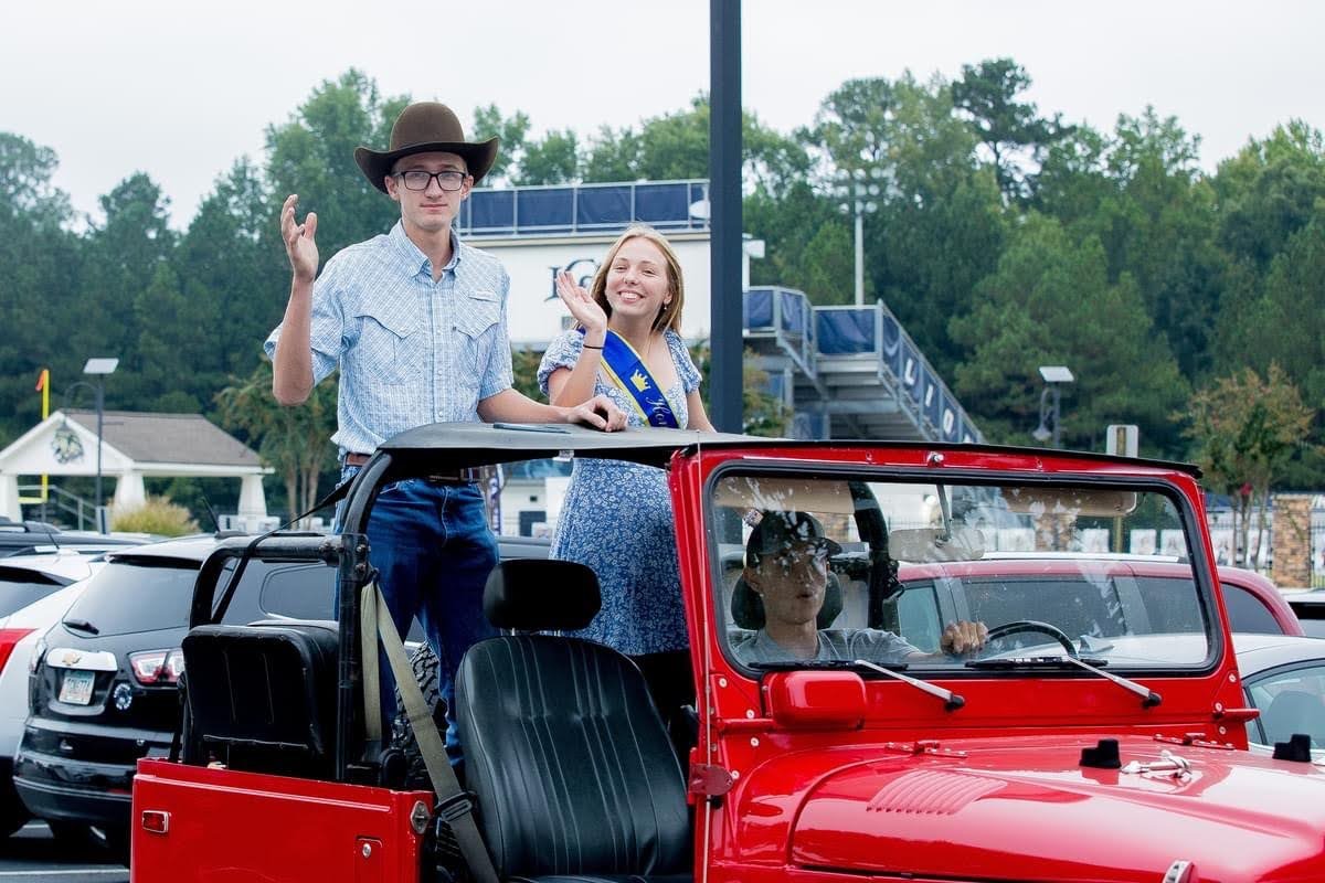 A man in a cowboy hat and a woman wearing a sash wave from the back of a red convertible jeep, with a sports field visible in the background.