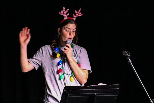 A person wearing a festive outfit with reindeer antlers and colorful lights around their neck is speaking into a microphone on stage.