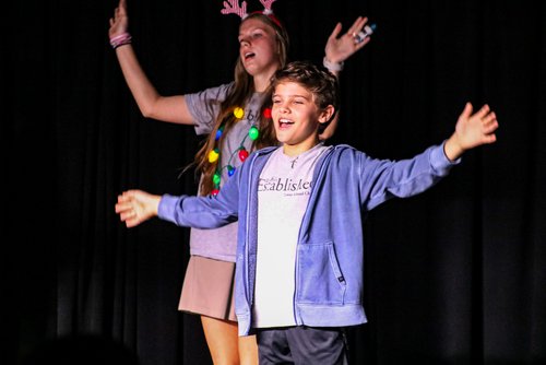 A young boy and girl perform on stage, with the girl wearing festive decorations and the boy smiling broadly with arms outstretched, both appearing joyful in a theatrical setting.