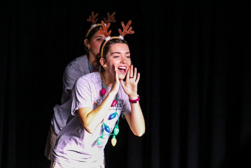 Two individuals perform on stage, one wearing reindeer antlers and colorful decorations, while the other stands behind, creating a playful scene.