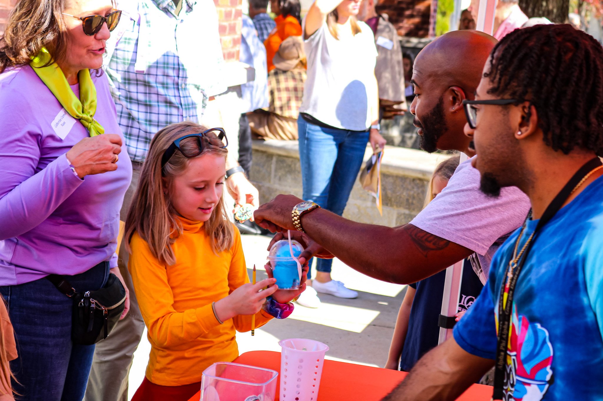 A young girl in a yellow shirt is receiving a colorful drink from a man at a table, while adults observe and engage in conversation in a lively outdoor setting.