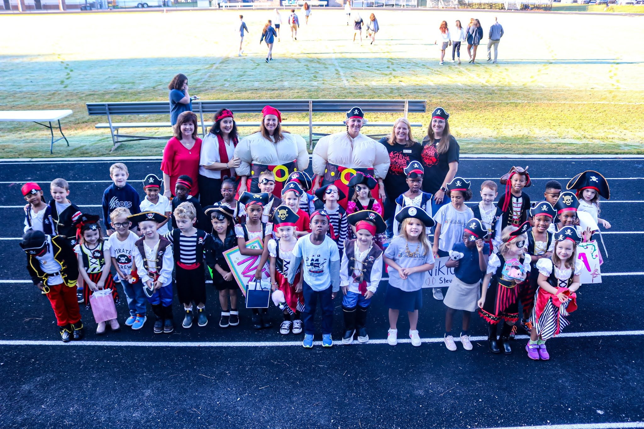 A large group of children dressed in pirate costumes, posing together on a sports field, with a few adults also in costume behind them.