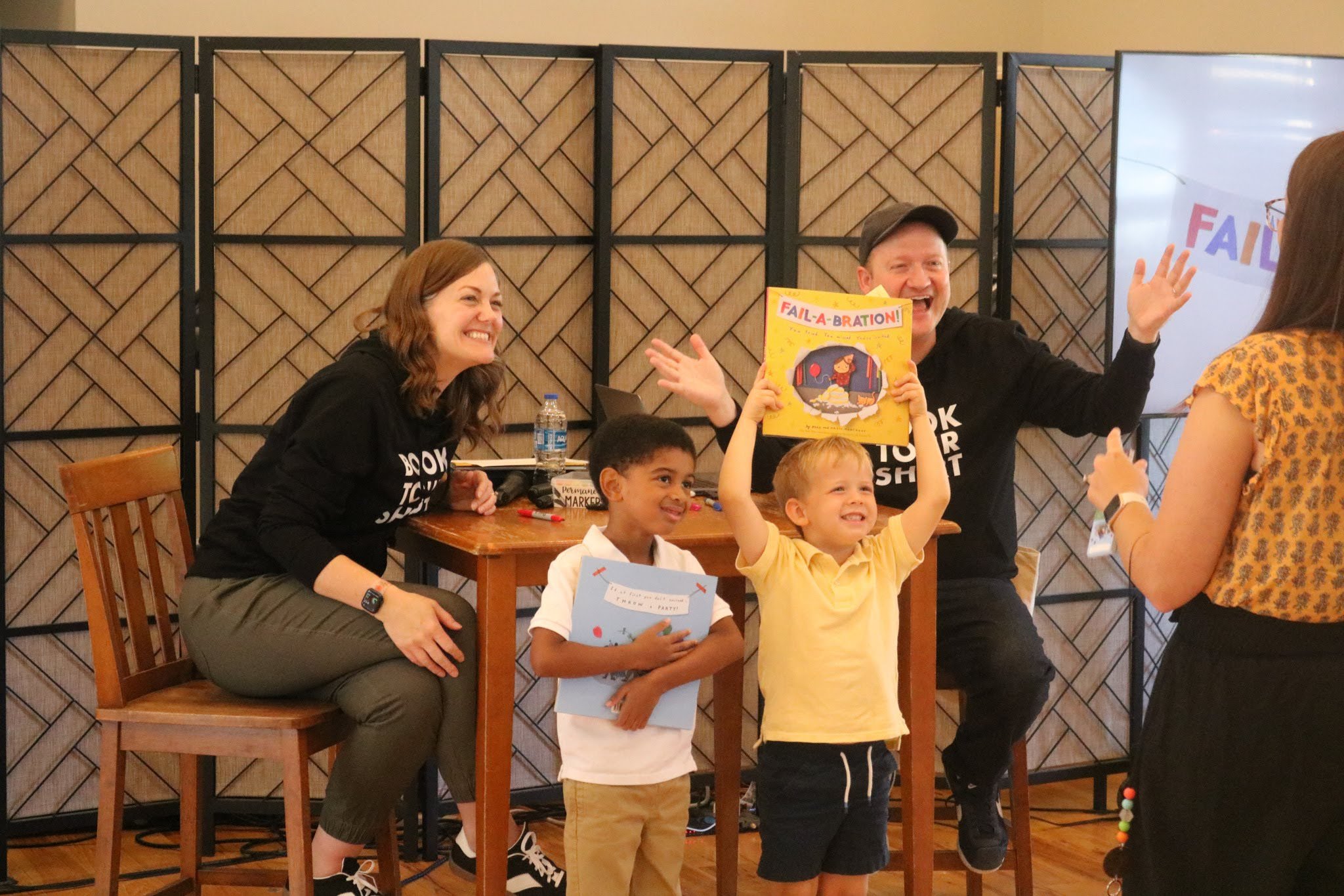 A lively scene featuring two children holding books, with one child raising a book high, while two adults seated nearby smile and engage with them during a reading event.