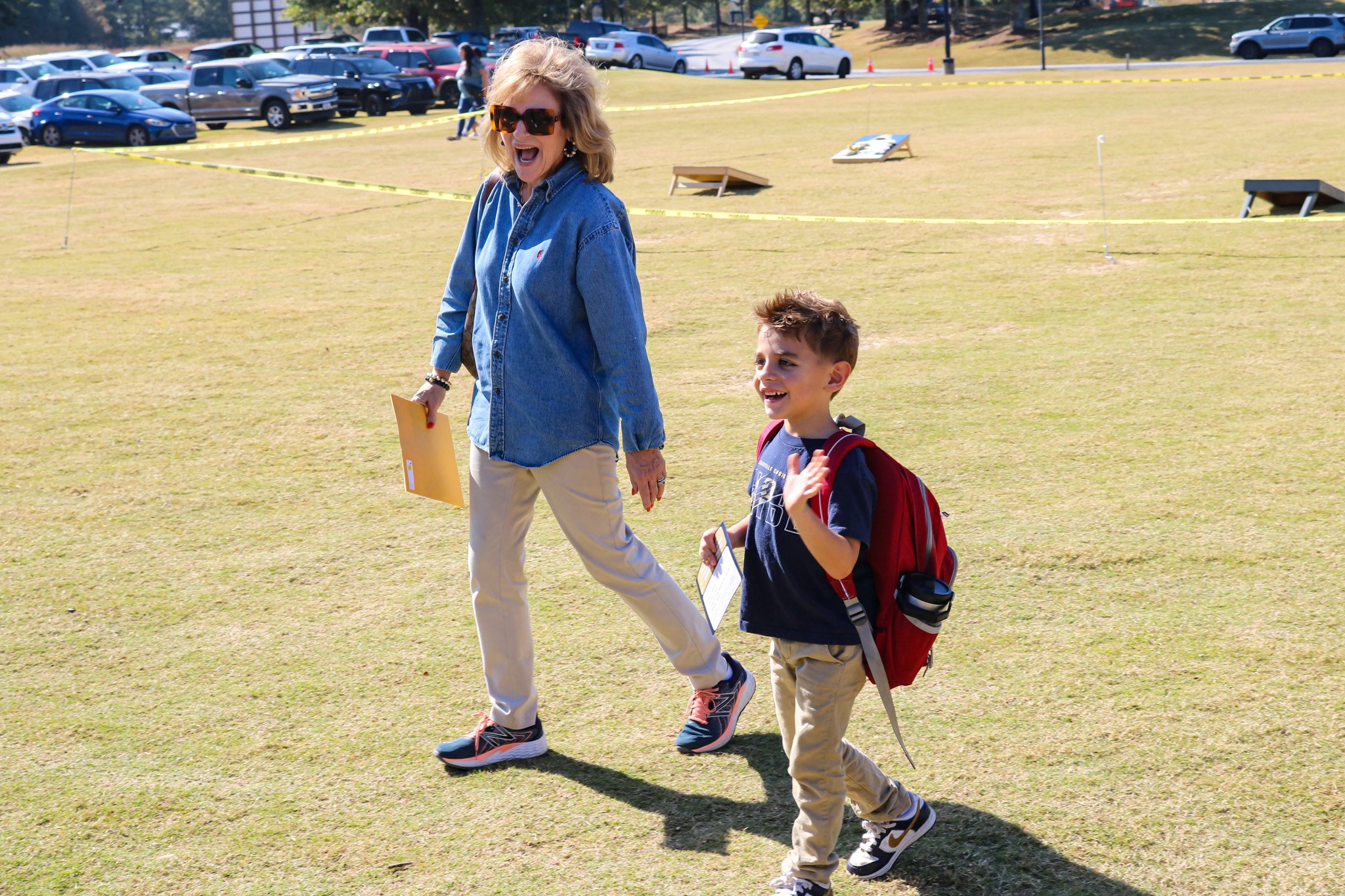 A woman and a young boy walk together across a grassy field, with the woman holding a clipboard and the boy carrying a backpack.