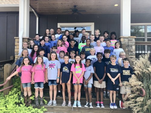 A large group of children and teenagers stands together on a porch, smiling for the camera, wearing a variety of colorful t-shirts and shorts.