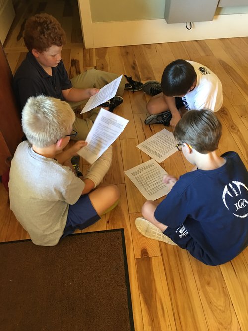 Four children sitting on a wooden floor, each reading from sheets of paper spread out in front of them.