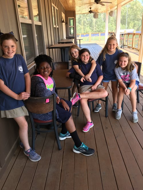 A group of six children, wearing matching blue shirts and shorts, are sitting on a wooden deck, smiling and posing for the camera.