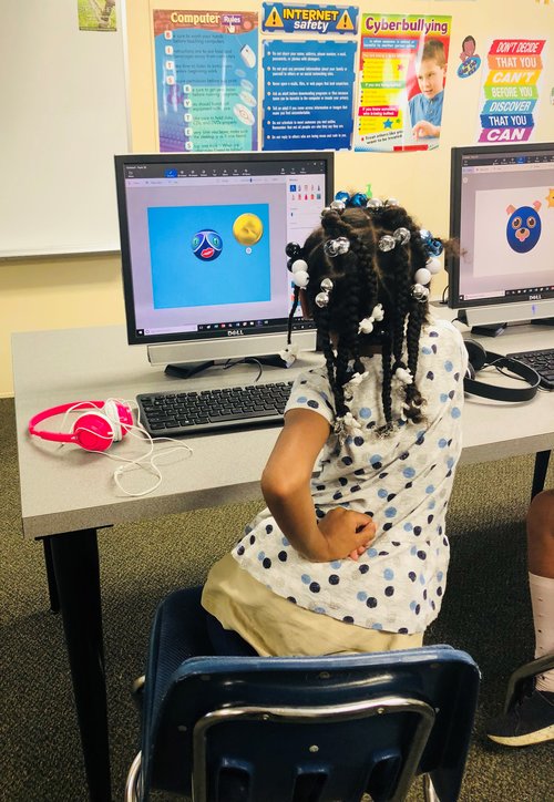 A young girl with braided hair sits at a computer in a classroom, looking at a colorful screen displaying animated images, while wearing headphones around her neck.