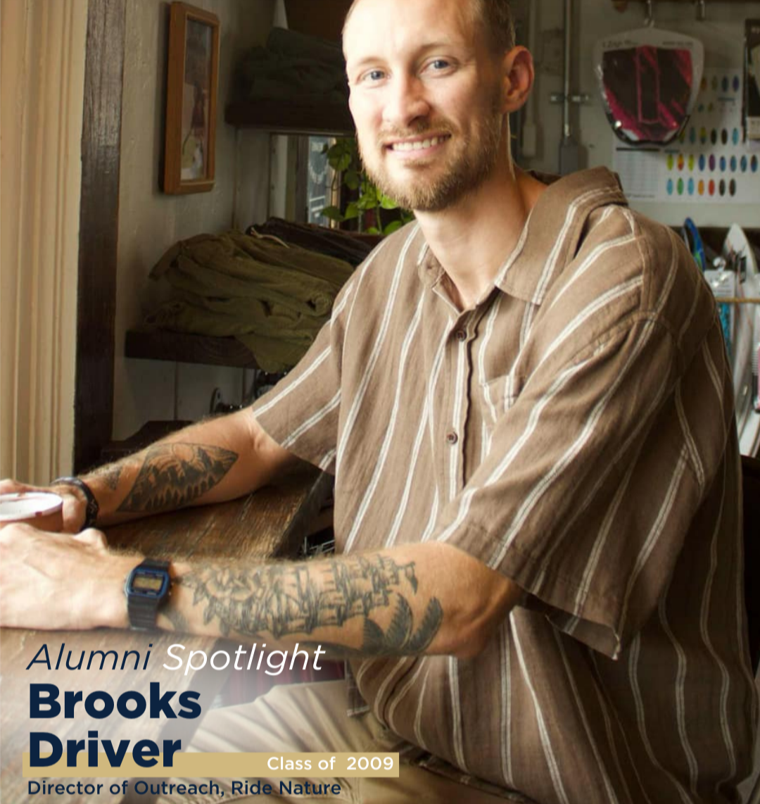 A smiling man with tattoos, wearing a striped shirt, sits at a table in a well-lit room filled with various items, promoting an alumni spotlight feature.
