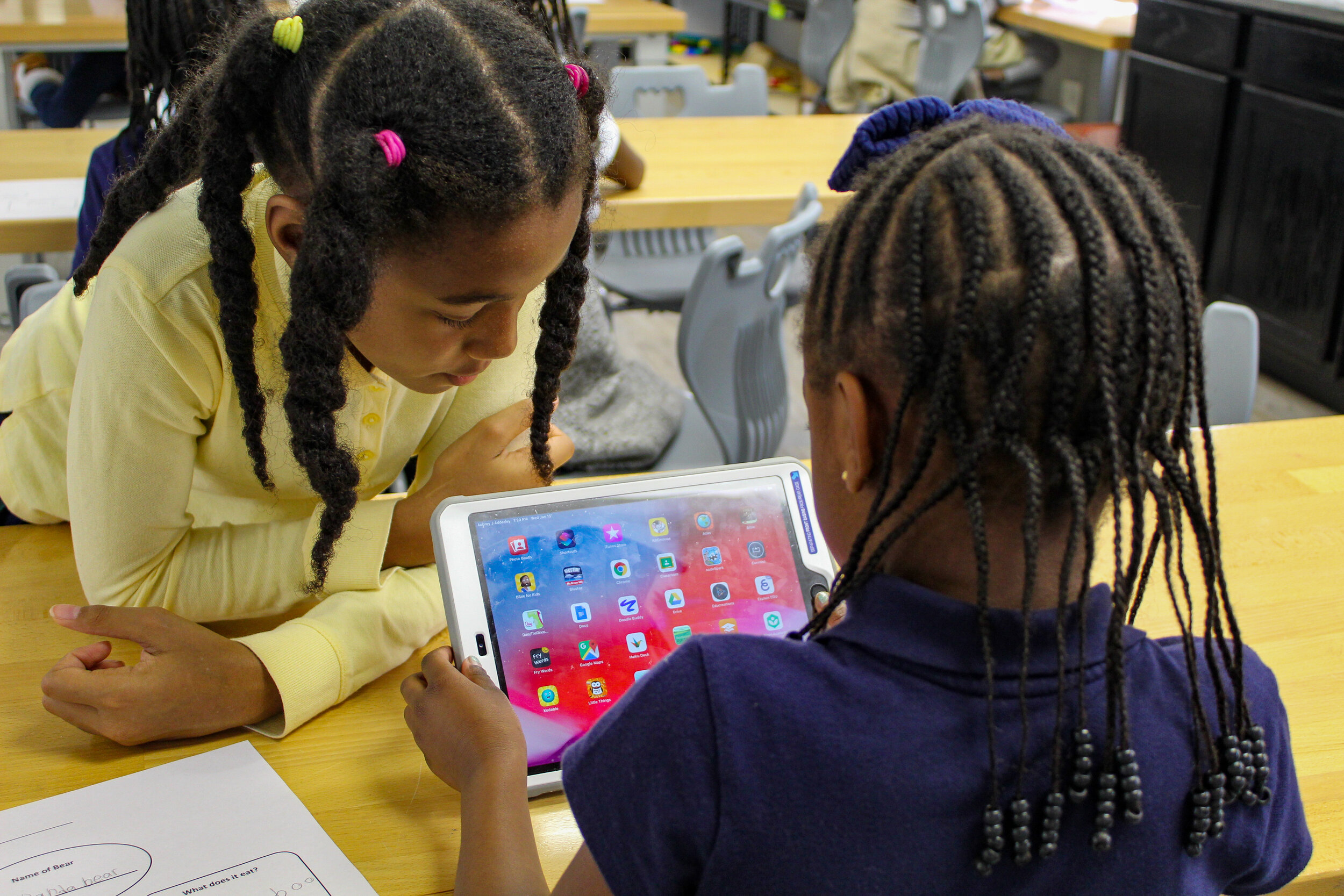 Two young girls are sitting at a table, closely examining a tablet displaying colorful app icons, with one girl leaning in to get a better view.