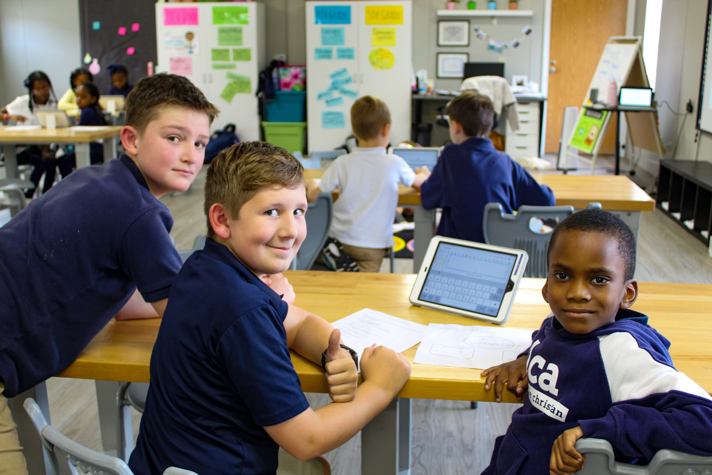 Three children are seated at a table in a classroom, smiling and looking at the camera, while other students work in the background with colorful posters on the walls.