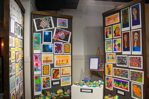 An art exhibition featuring colorful framed artworks displayed on walls, with a variety of paintings and sculptures arranged on a table in the foreground.