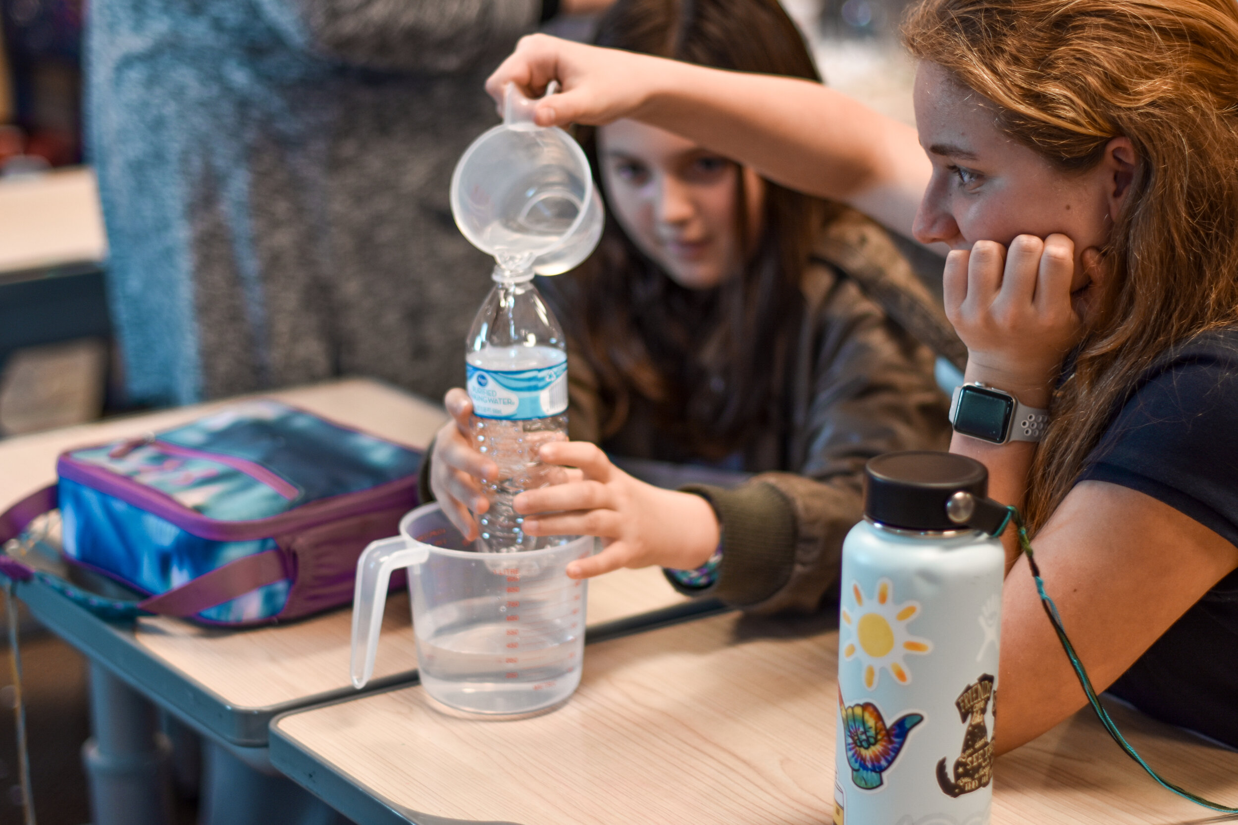Two children are engaged in a science experiment, one pouring water from a measuring cup into a plastic bottle while the other observes thoughtfully.