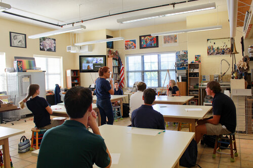 A classroom setting with a teacher addressing students seated at tables, surrounded by art supplies and educational materials.