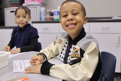 A smiling boy in a colorful jacket sits at a table, writing with a crayon, while a girl in the background also engages in an activity.