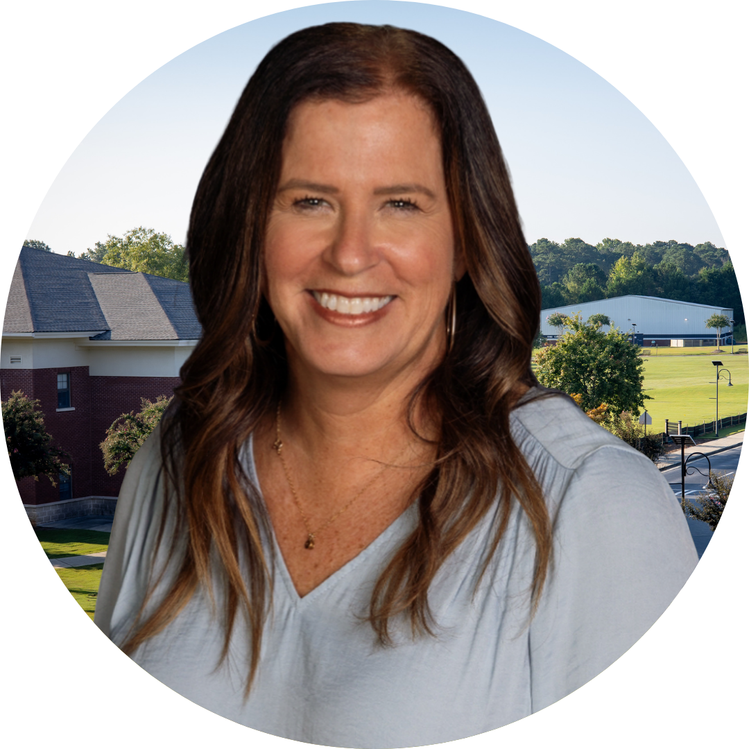 A smiling woman with long brown hair, wearing a light blue blouse, stands in front of a blurred building background.