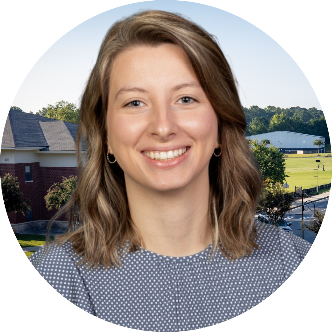 A smiling woman with long, wavy blonde hair is seated in front of a classroom setting, which includes wooden desks and a whiteboard in the background.