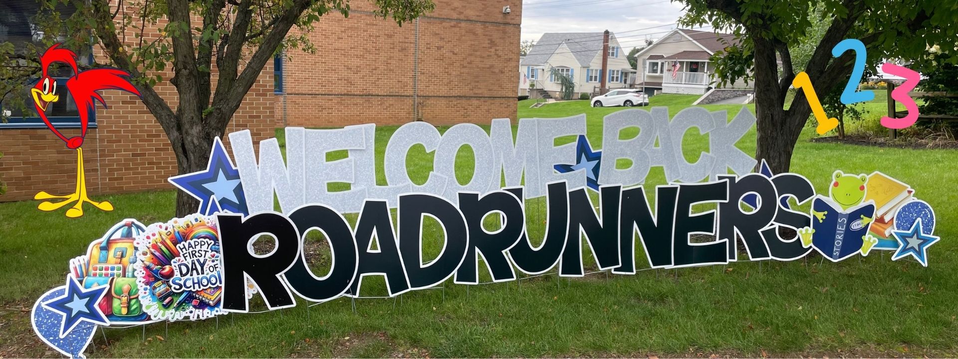 A colorful welcome sign for "Roadrunners" is displayed on a lawn with trees. The background shows a building and houses.