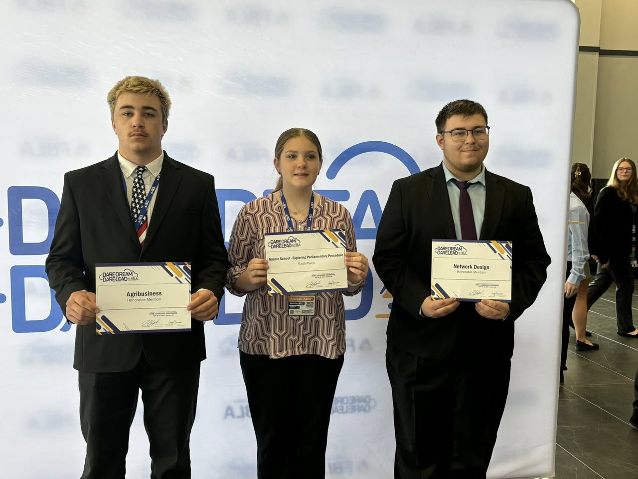 Three people stand together, holding certificates, with a festive banner in the background.