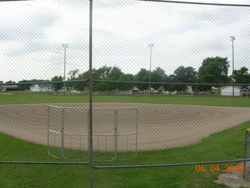  A baseball field surrounded by a fence, showcasing a neatly groomed diamond and vibrant green grass under sunny weather.