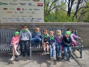 A group of children wearing green bandanas sits together on a bench, smiling and enjoying their time outdoors.