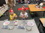 Two children in party hats sit at a table, joyfully painting with colorful supplies spread around them.