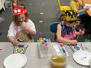 Two children wearing crowns sit at a table, surrounded by paint and paper, engaged in a creative art activity.