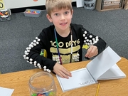 A young boy sits at a table, focused on a book, with pencils scattered around him.