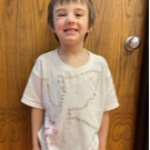 A young boy wearing a shirt with text printed on it, smiling and standing outdoors.