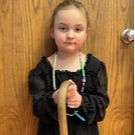 A young girl in a black dress stands holding a cane, looking confidently at the camera.