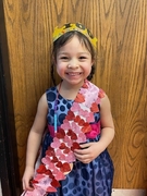 A young girl in a sash holds a heart-shaped paper, smiling brightly as she poses for the camera.