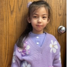 A young girl in a purple sweater and skirt smiles while standing outdoors.