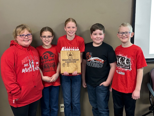 Five children in red shirts proudly hold a plaque, smiling at the camera in a bright outdoor setting.