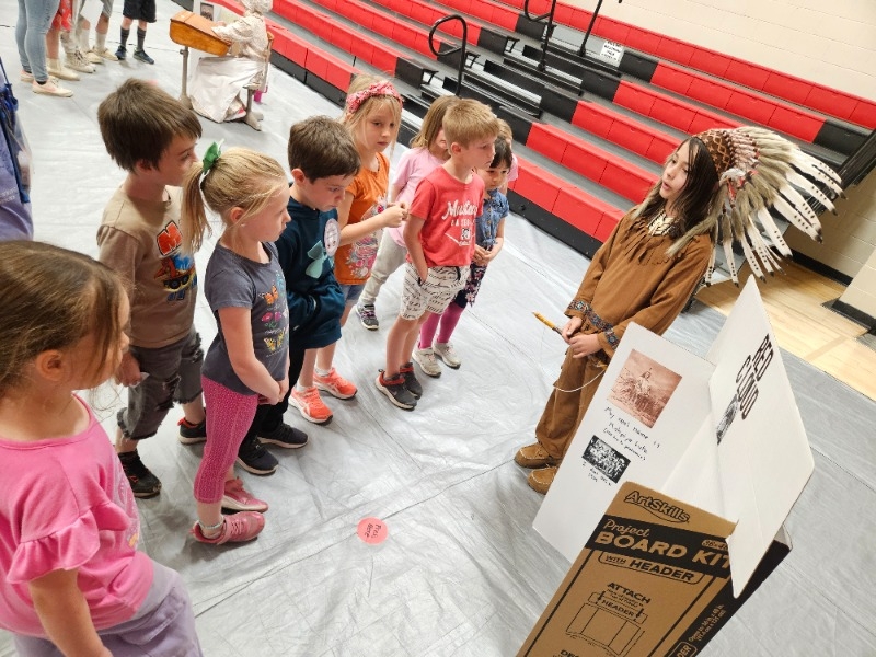 Children in Native American costumes stand in line, eagerly waiting to see a cardboard box in front of them.