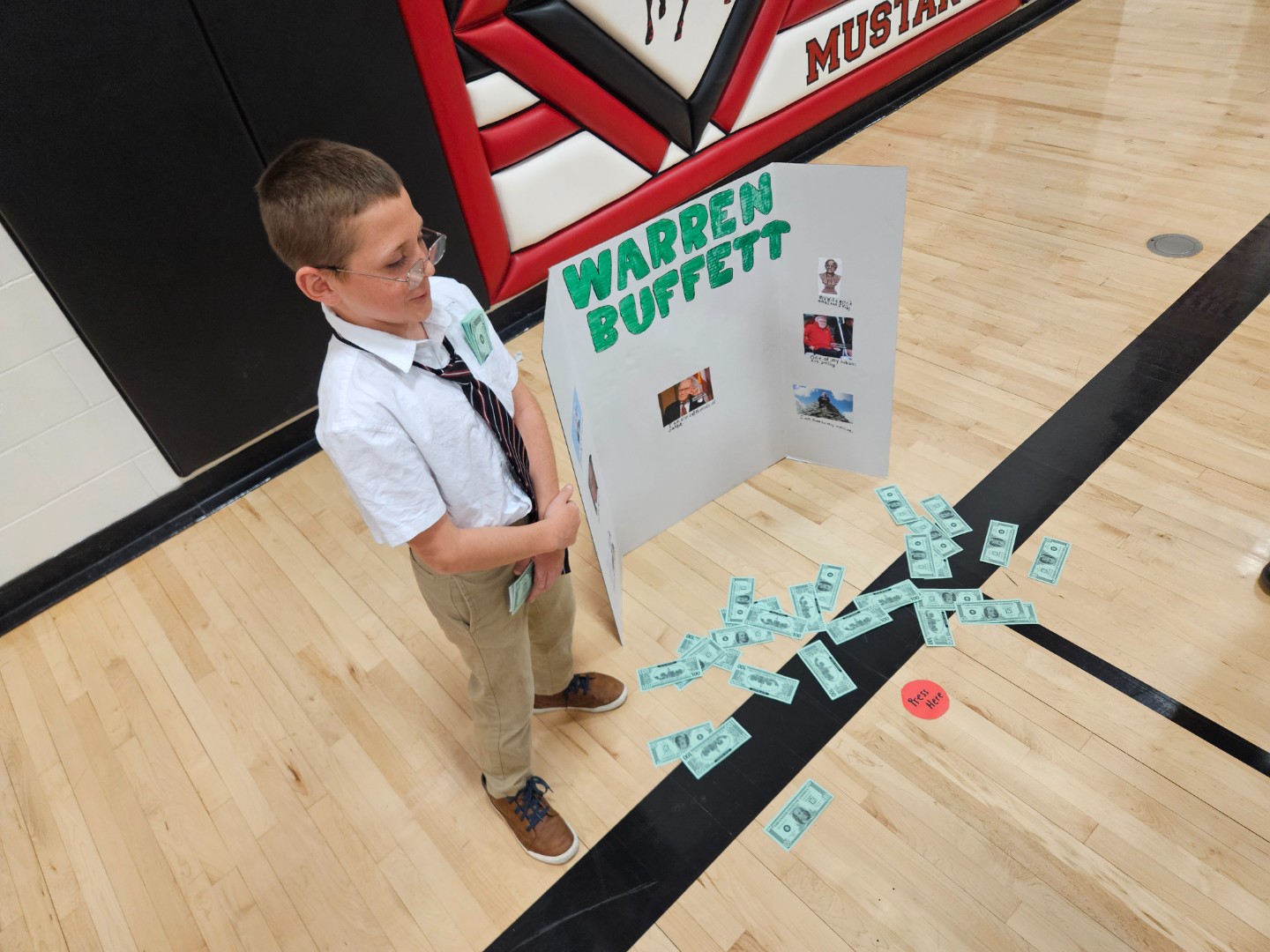 A boy in a tie stands beside a poster board displaying various denominations of money.