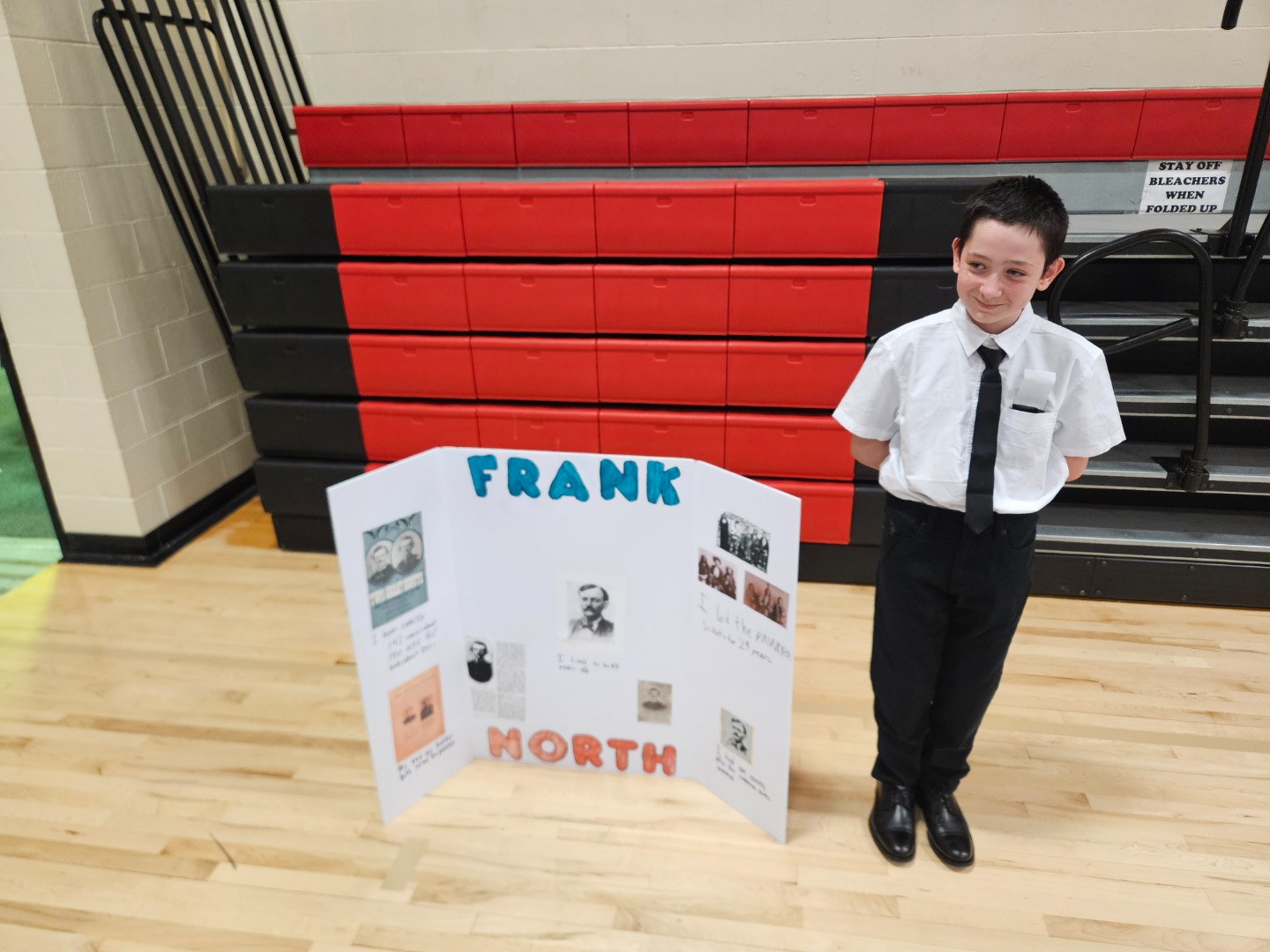 A young boy stands beside a poster board displaying various pictures, smiling and pointing at one of the images.