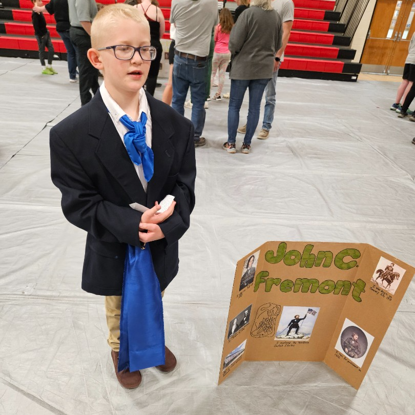 A boy in a suit and tie stands beside a poster board, smiling confidently at the camera.