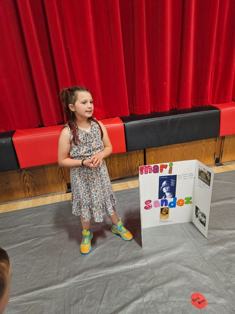 A young girl stands proudly in front of a colorful poster board displaying various drawings and information.