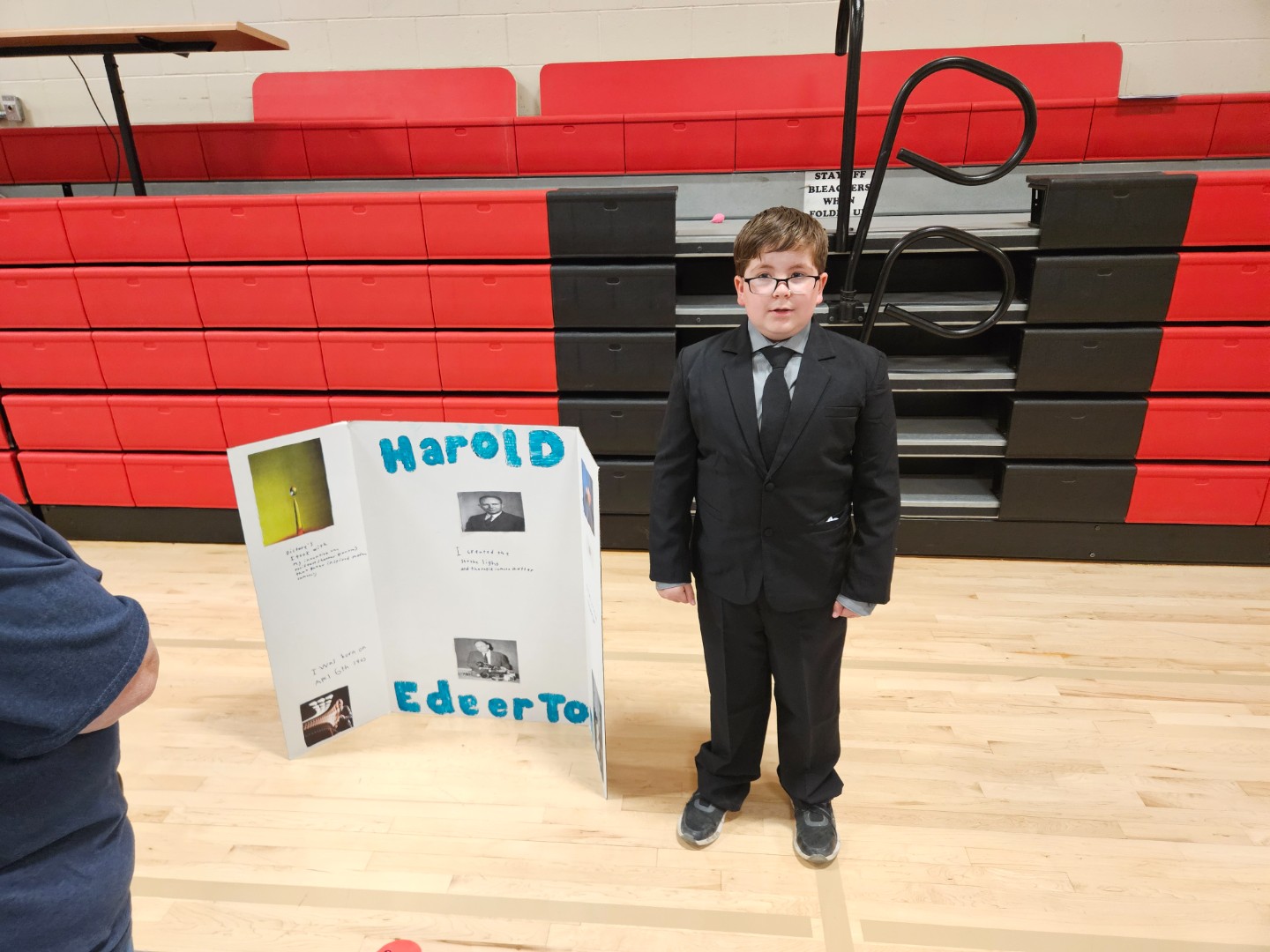 A boy in a suit stands beside a poster board, smiling and looking at the camera.