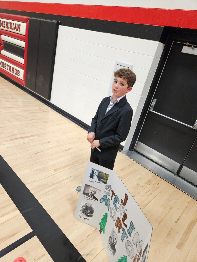 A boy in a suit stands confidently in front of a colorful poster board displaying various educational materials.