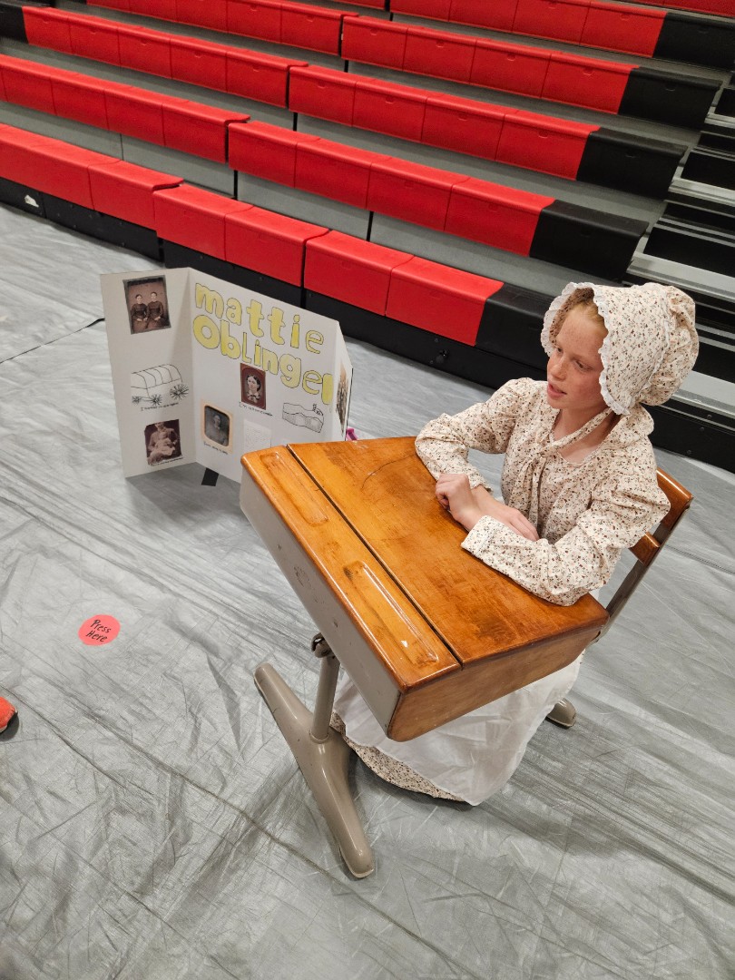 Girl in historic dress at desk next to "Mattie Oblinger" display. Classroom setting.