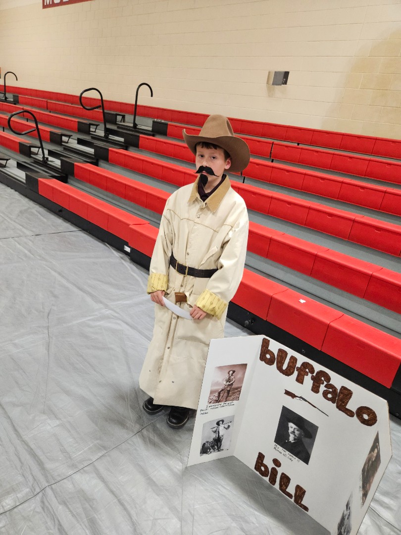 A young boy dressed as a cowboy stands proudly in front of a wooden sign, smiling at the camera.