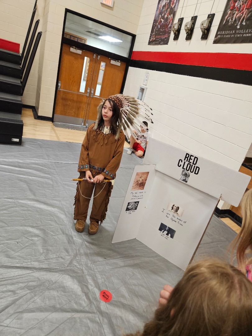 A young girl in a Native American costume stands proudly in front of a colorful sign, smiling at the camera.