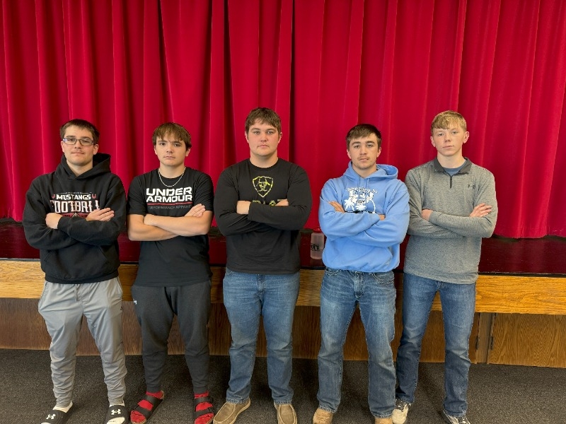 Five young men pose together in front of a vibrant red curtain, smiling and dressed casually.